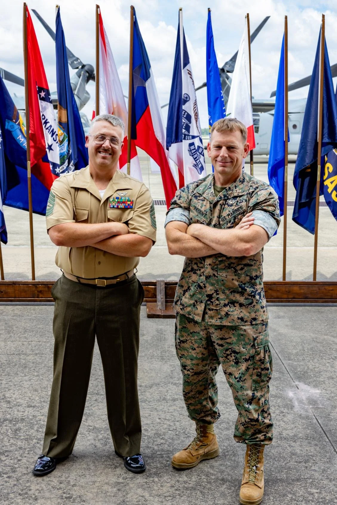 Two military men standing with arms crossed in front of a row of flags at an airfield. The man on the left is in a tan uniform with various medals, and the man on the right is in camouflage military gear.