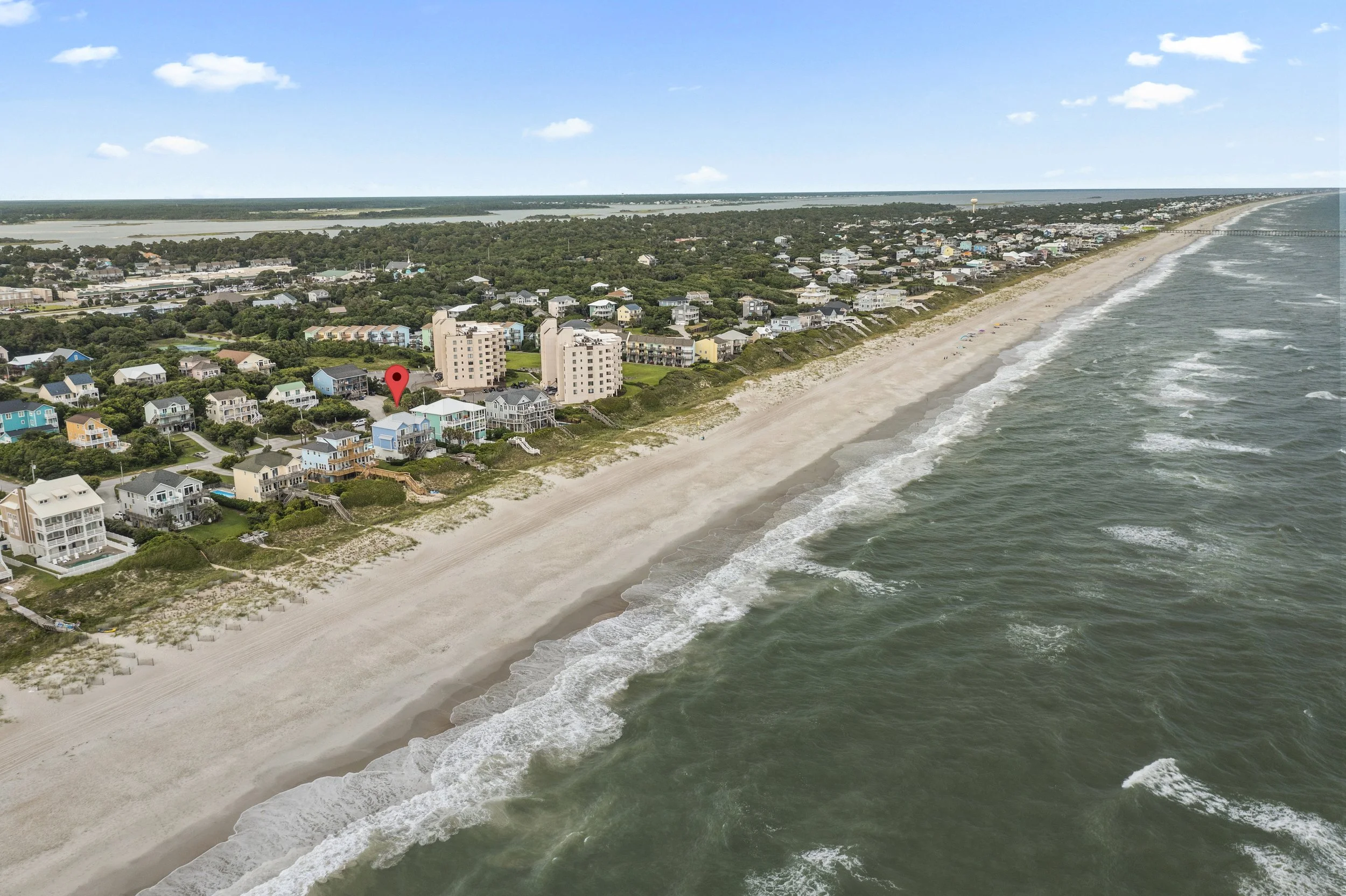 Aerial view of a beach with shoreline, ocean waves, and colorful houses near the coast, with a forested area and town in the background under a blue sky with scattered clouds.