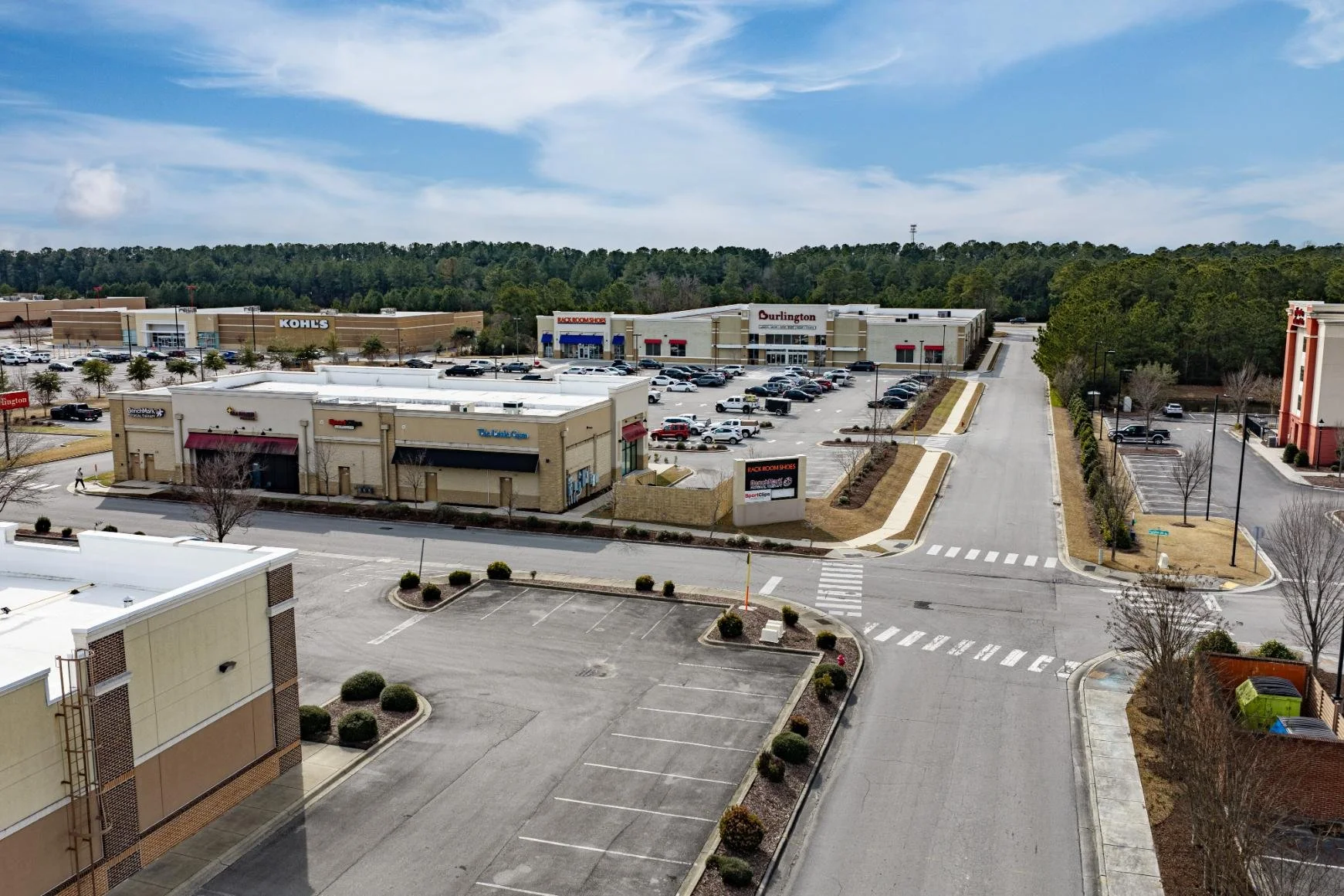 A shopping plaza with several retail stores including Kohl's, Dick's Sporting Goods, and Burlington, with a parking lot and a wooded area in the background under a partly cloudy sky.