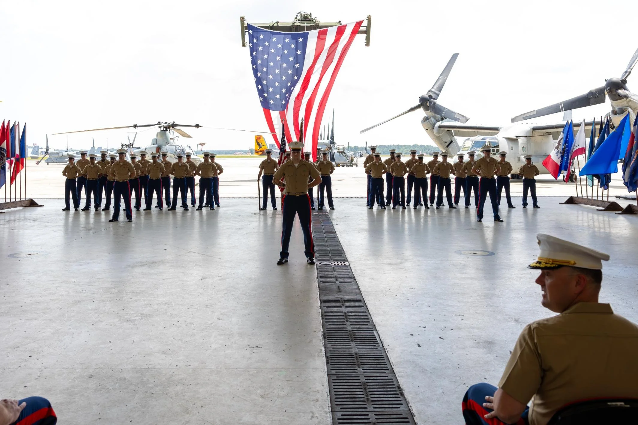 Military personnel standing in formation with an American flag hanging from a large aircraft. Several helicopters and aircraft are visible on the tarmac during an airbase setting.