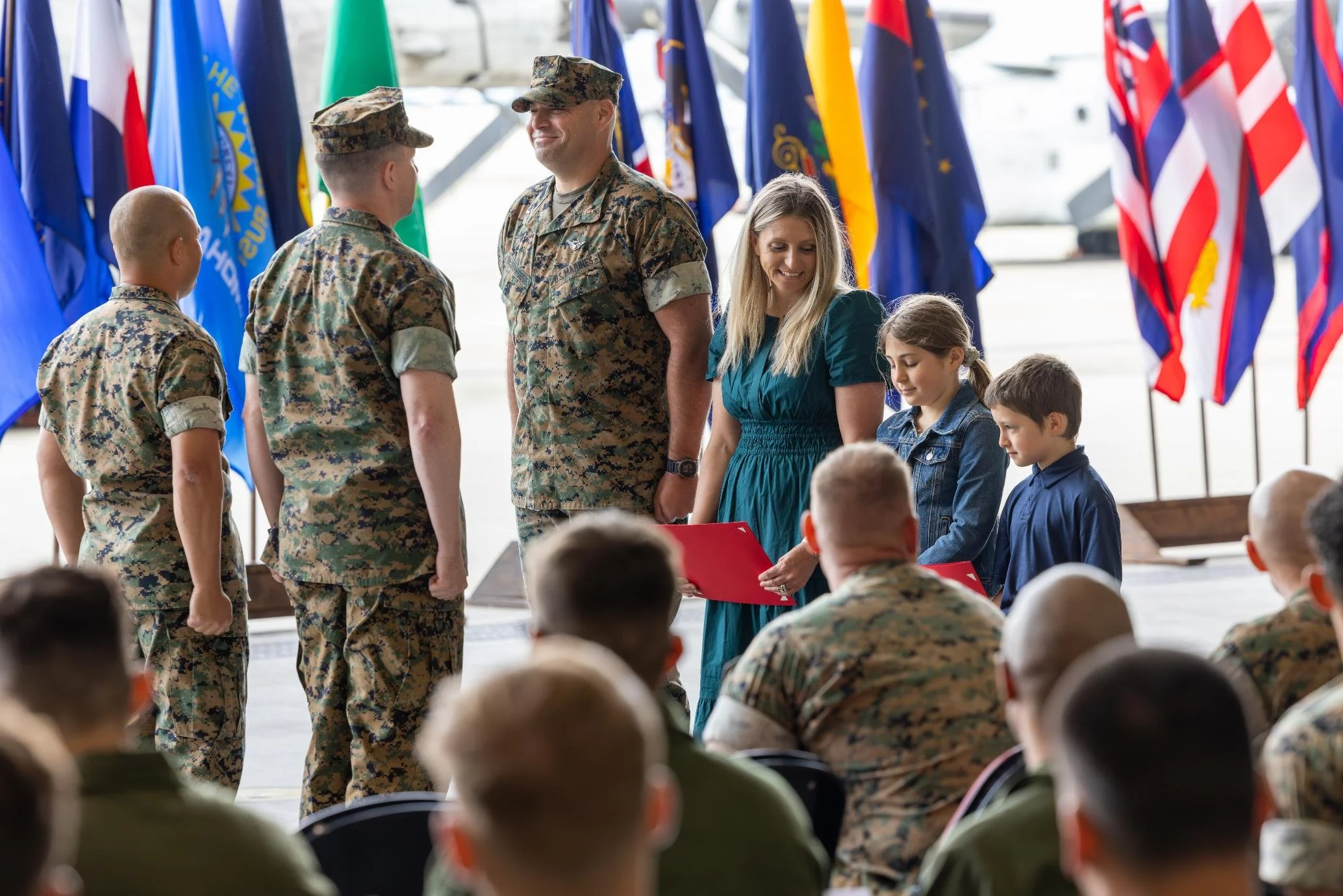 U.S. military personnel in uniform standing in line while two women, including one in a blue dress, present them with certificates or awards during a ceremony with multiple flags in the background.