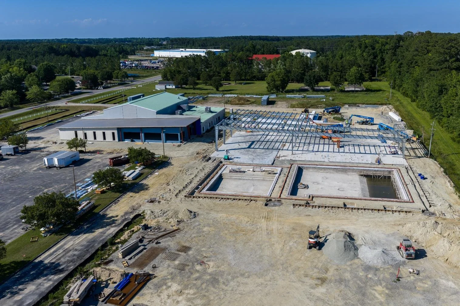 Aerial view of a construction site with steel framework, concrete pools, and a partially built building, surrounded by trees and parking lots.