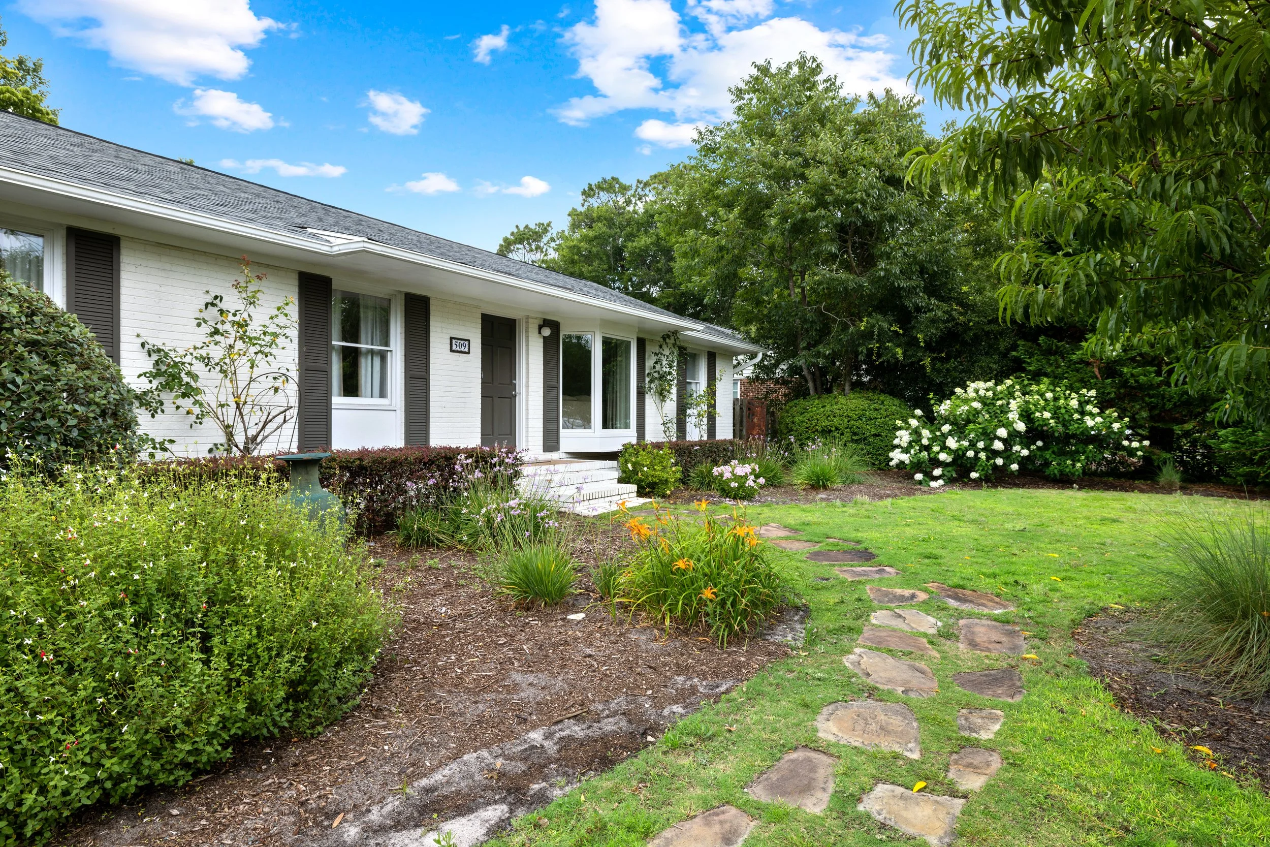 Front yard of a white house with a gray roof, surrounded by green trees and colorful flowers, walking stones on a grass lawn leading to the front door.
