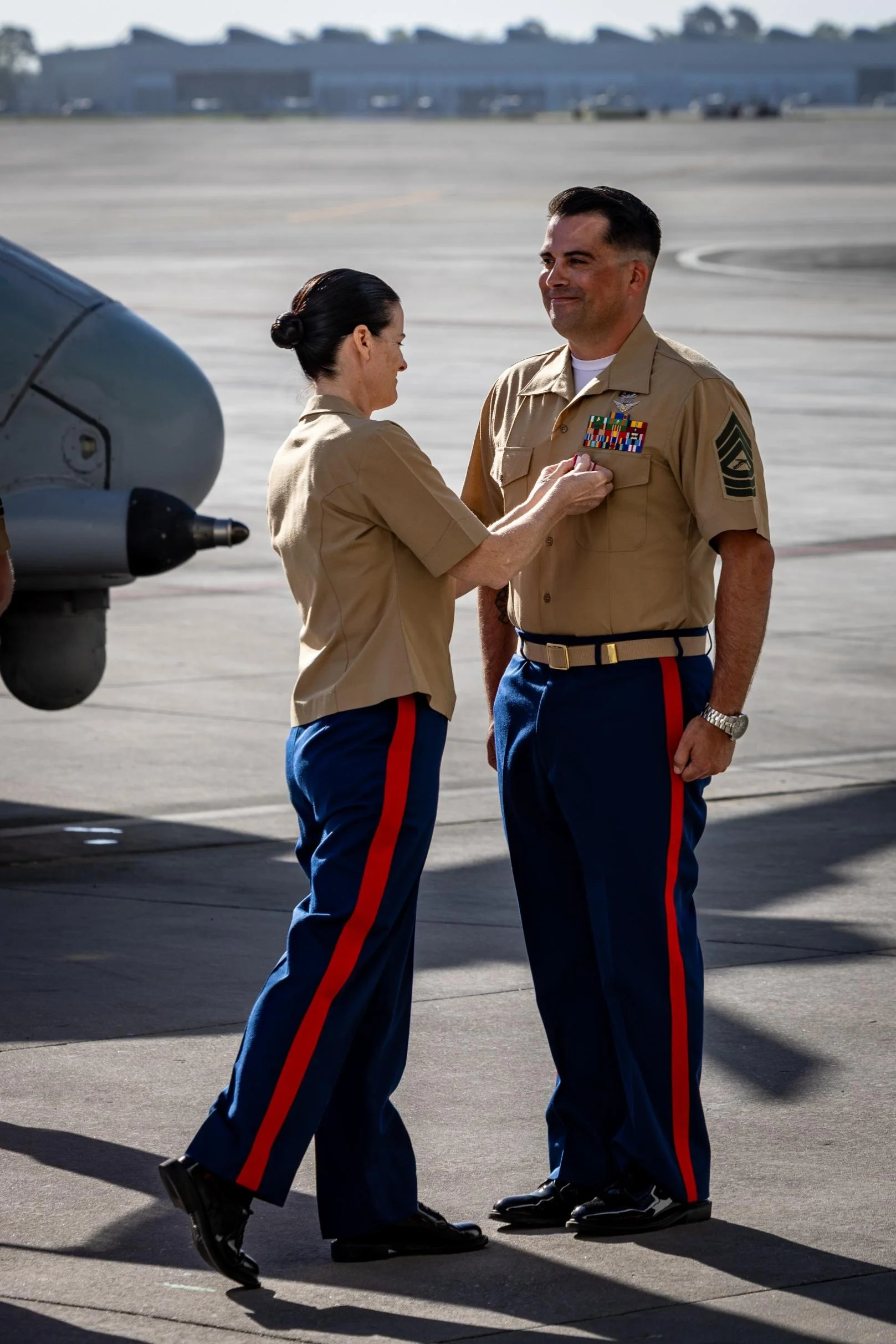 A woman is pinning a medal or ribbon onto a male military officer's uniform at an airfield, both are dressed in military dress uniforms. The woman has dark hair in a bun, and the officer has short dark hair and a wristwatch.
