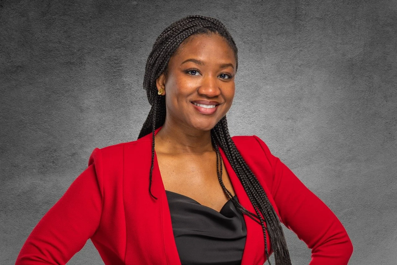 A woman smiling, wearing a red blazer and black top, with braided hair and gold earrings, standing against a gray textured background.