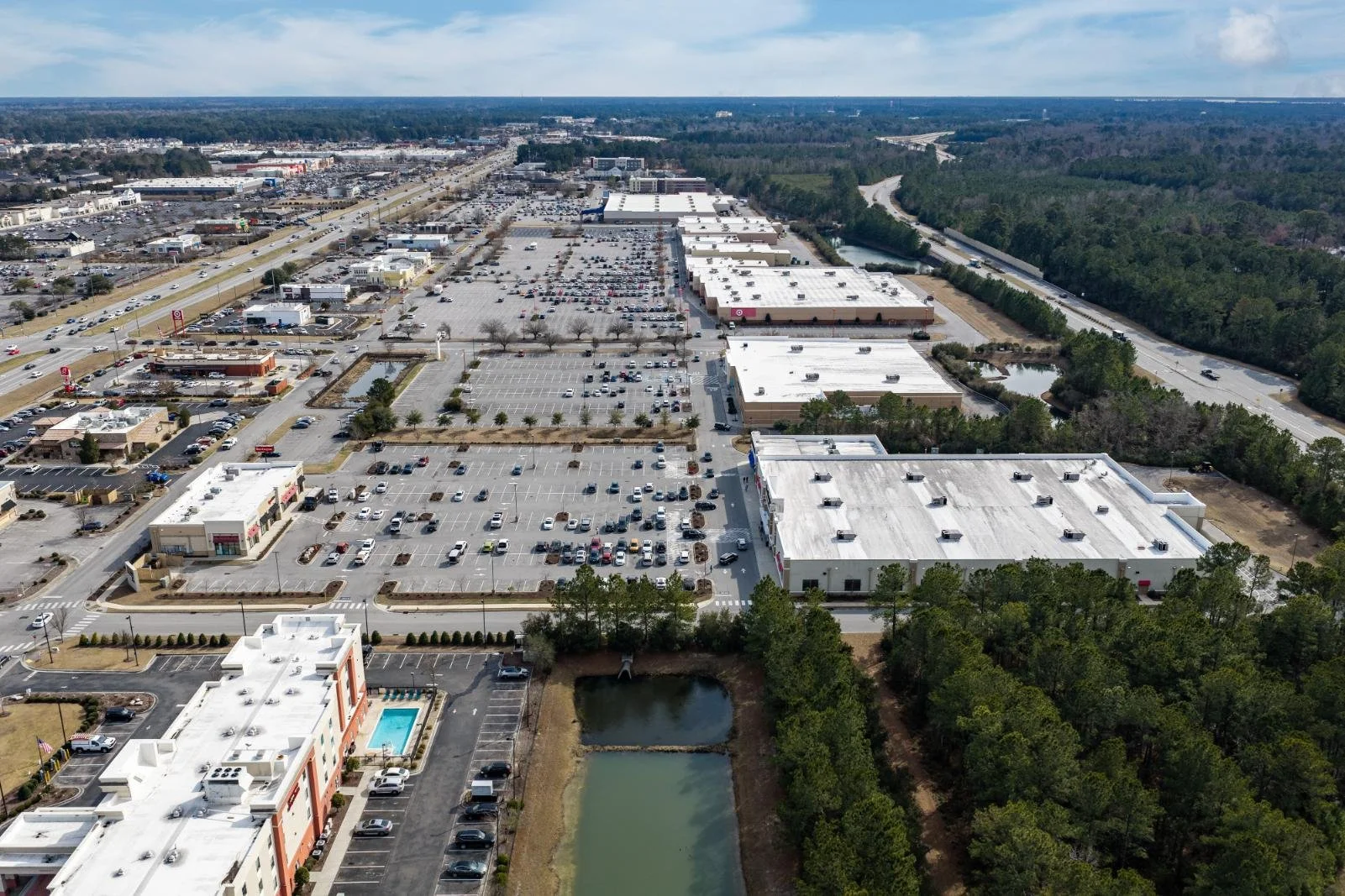 Aerial view of a shopping center with large parking lots, retail stores, and surrounding roads, with a wooded area and water bodies on the right side.