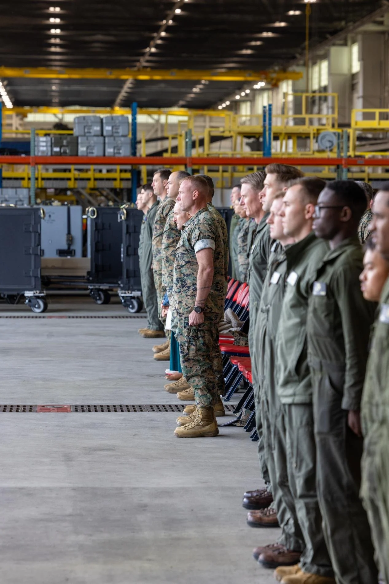 Military personnel standing in formation inside a warehouse or hangar with shelves and equipment in the background.