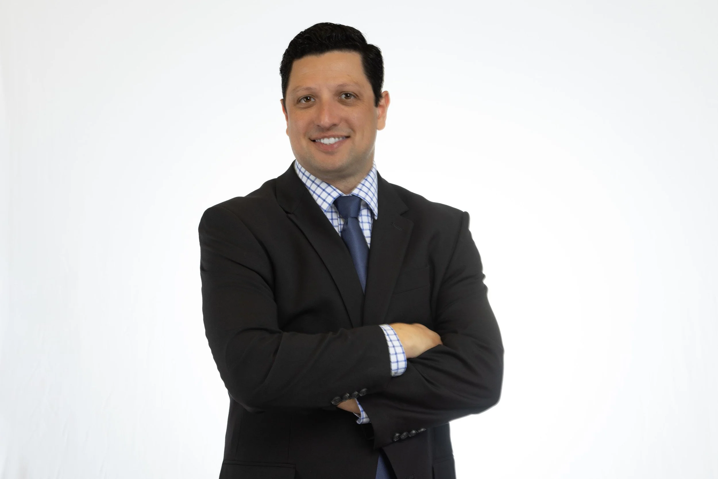 A man with dark hair wearing a black suit, blue tie, and checked shirt, standing with arms crossed against a plain white background.