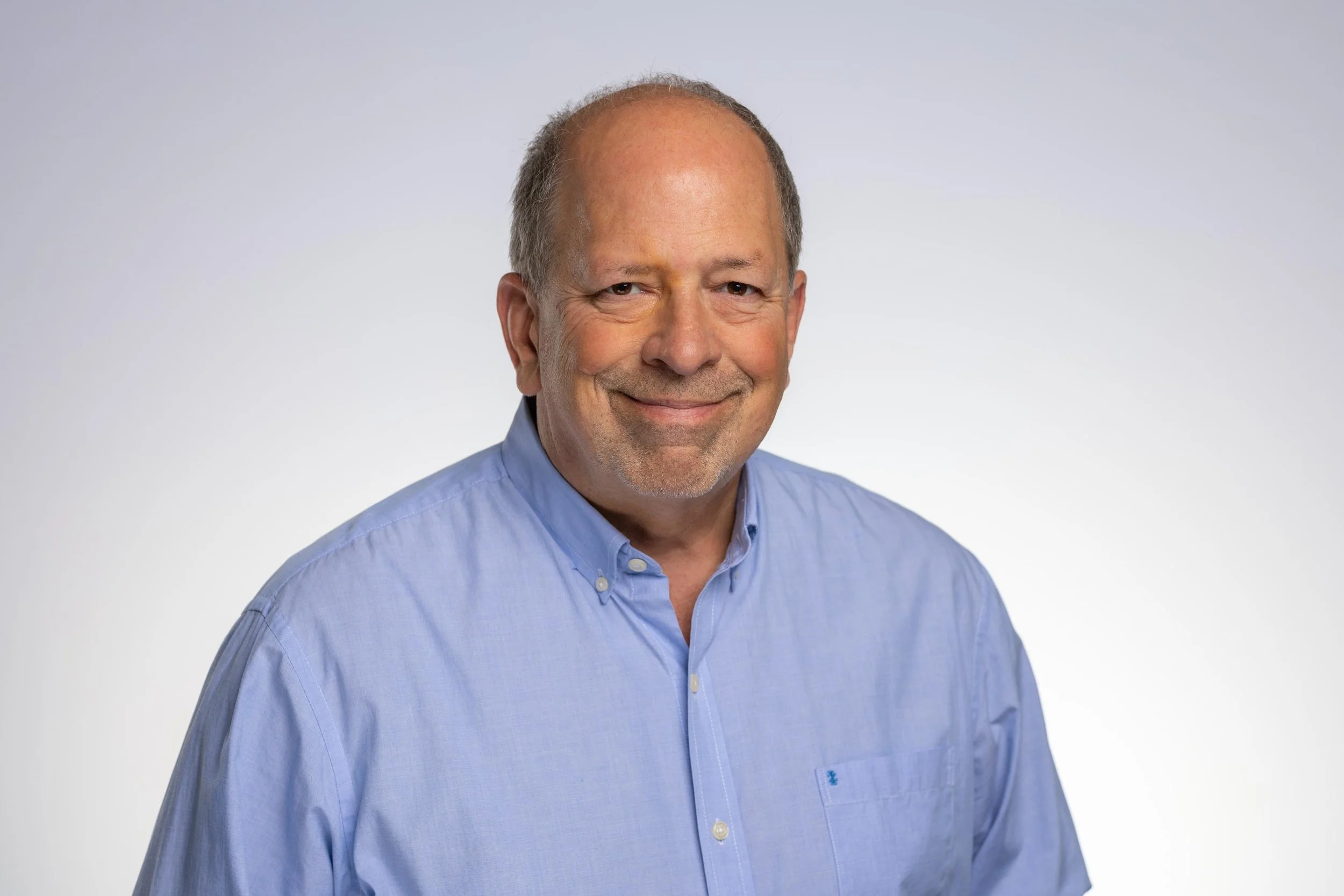 A middle-aged man with a smile, bald on top with gray hair on the sides, wearing a light blue button-up shirt, against a plain light background.