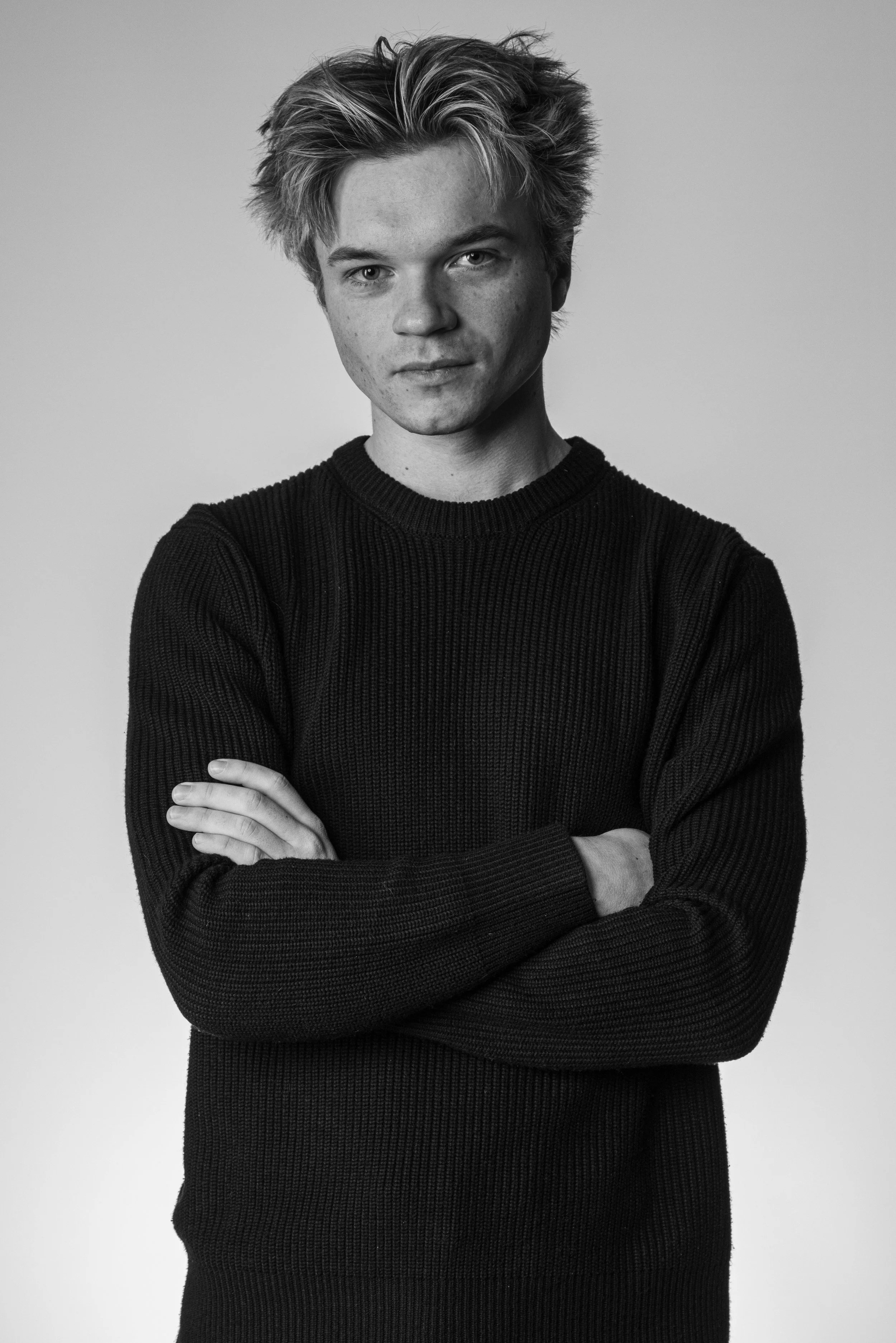 Black and white portrait of a young man with tousled hair, crossing his arms, wearing a dark, ribbed sweater, looking directly at the camera.