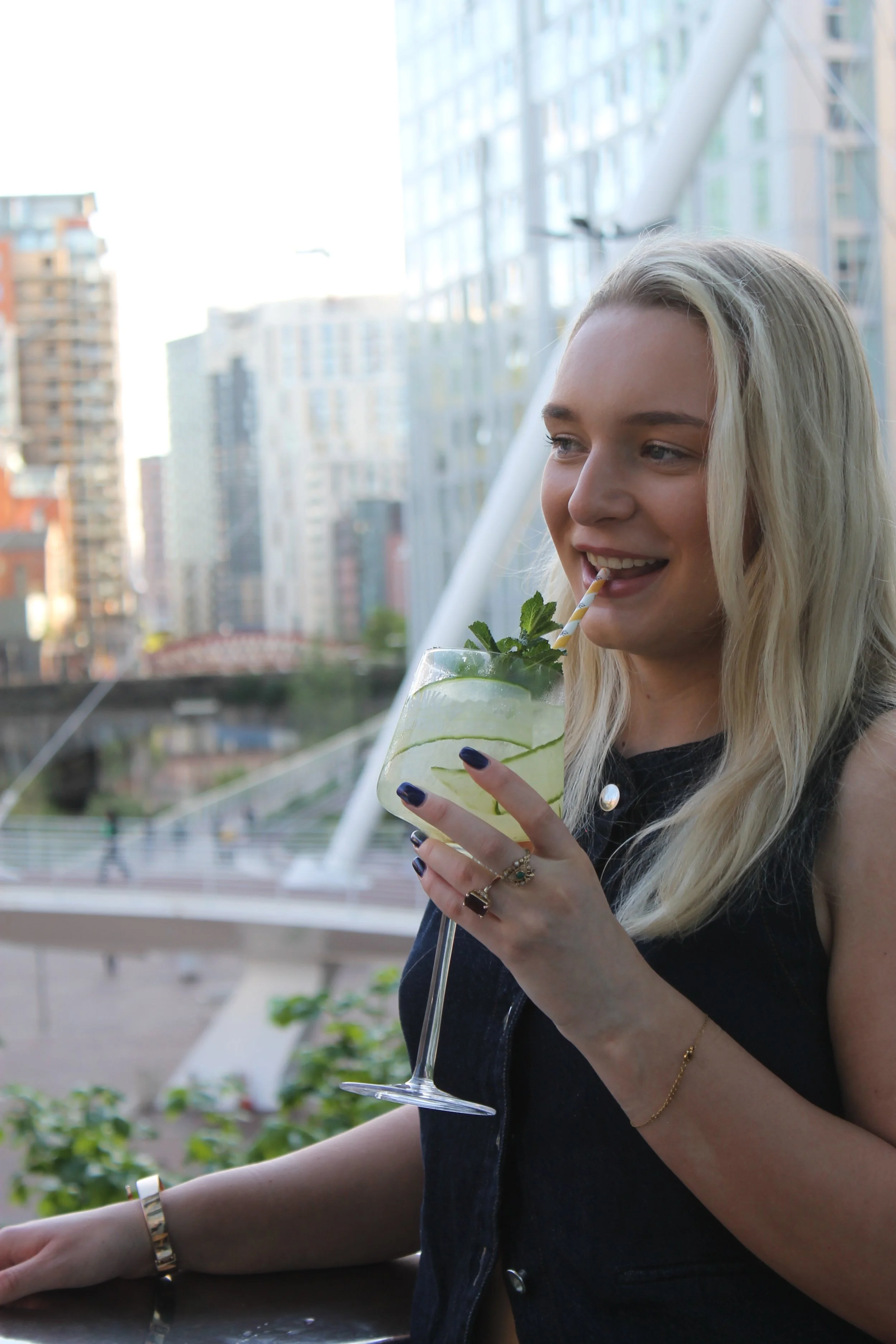 A young woman with blonde hair enjoying a lime cocktail with mint and lime slices on a balcony overlooking a city skyline.