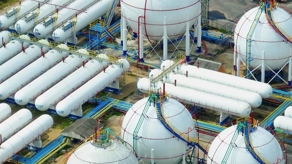 Industrial facility with large white spherical tanks, long white cylindrical tanks, and colorful piping and walkways.