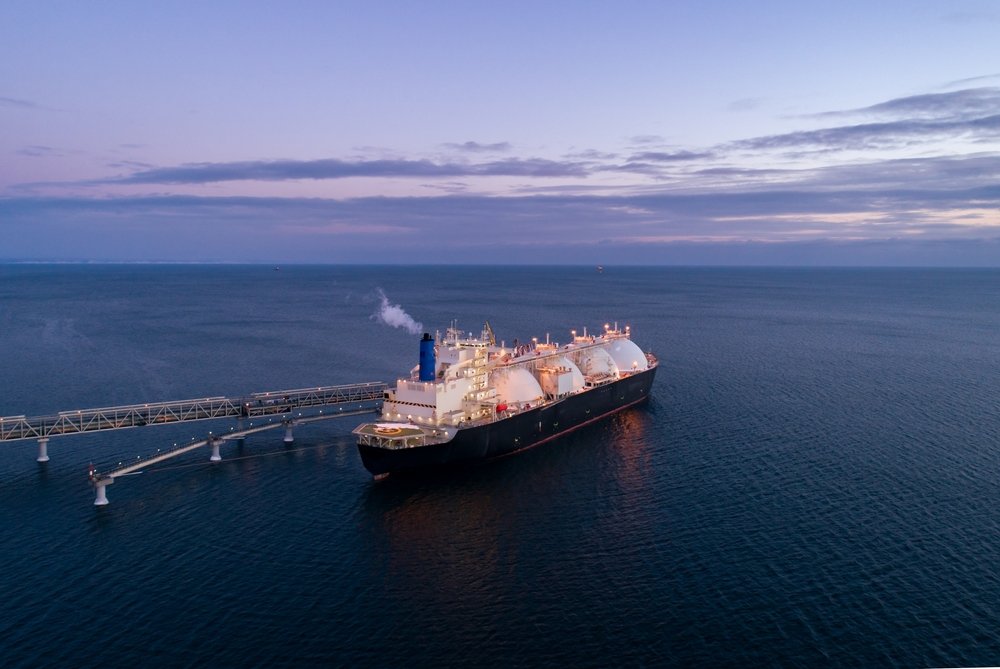 A large cargo ship, possibly an LNG tanker, sailing in open water near sunset or sunrise with a long dock or pipeline extending from the ship into the water.