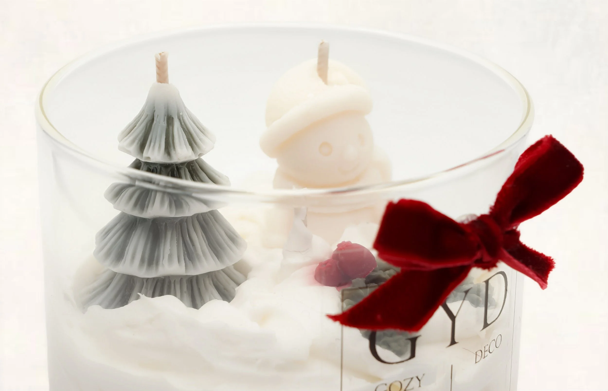 Decorative candle with a gray Christmas tree, white ornaments, small red berries, and a red bow in a clear glass container.