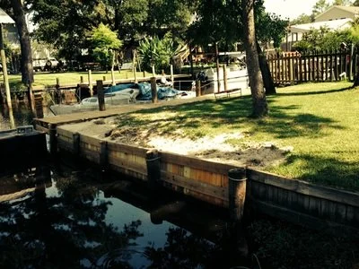 A backyard with a wooden dock extending over a canal, grassy yard, trees, and a boat docked by the canal.