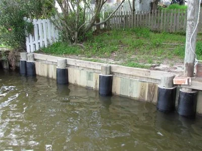 Waterfront with wooden and concrete retaining wall, black flotation devices, and a grassy area with trees and a white fence.