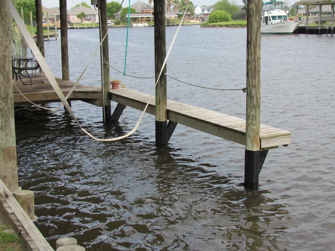 A wooden dock with a bench and ropes, extending over water in a residential neighborhood with boats and houses in the background.