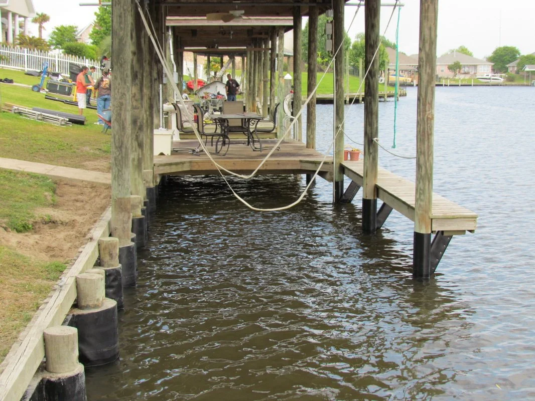 Wooden dock with furniture extending over a body of water, with people and boats visible in the background.