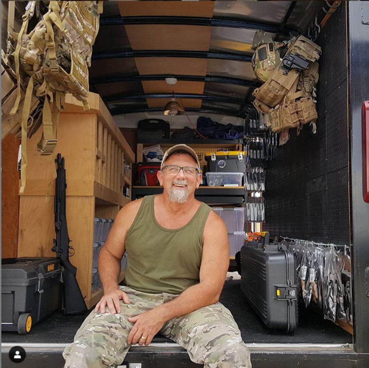 A smiling owner with a beard, wearing airsoft gear and camouflage pants, sitting inside an organized mobile airsoft gear storage trailer .