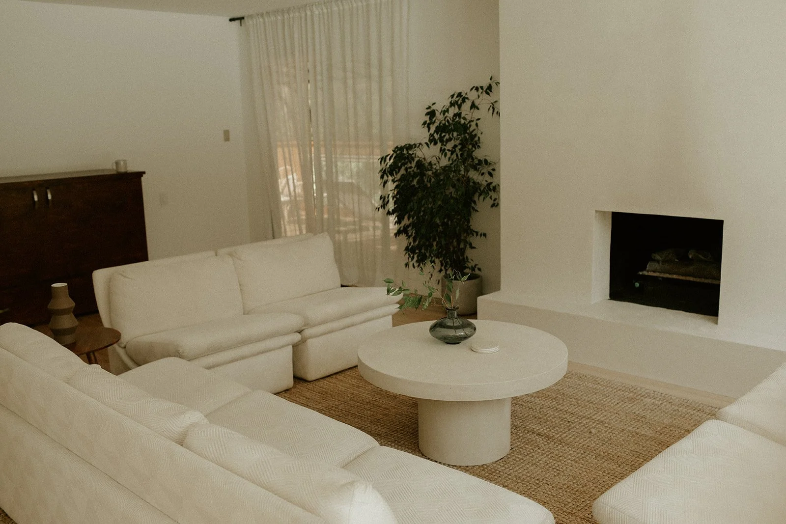 Living room with white sofas, a round coffee table with a decorative vase and small object, a potted plant, a wooden cabinet, and a fireplace, with a window covered by sheer curtains.