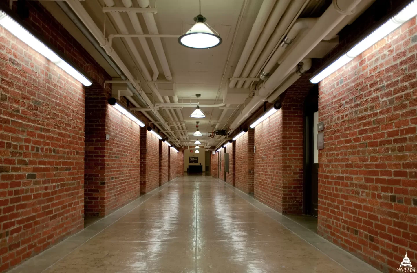 Long corridor with exposed brick walls, ceiling with visible pipes and ducts, bright overhead lighting, and a polished concrete floor.