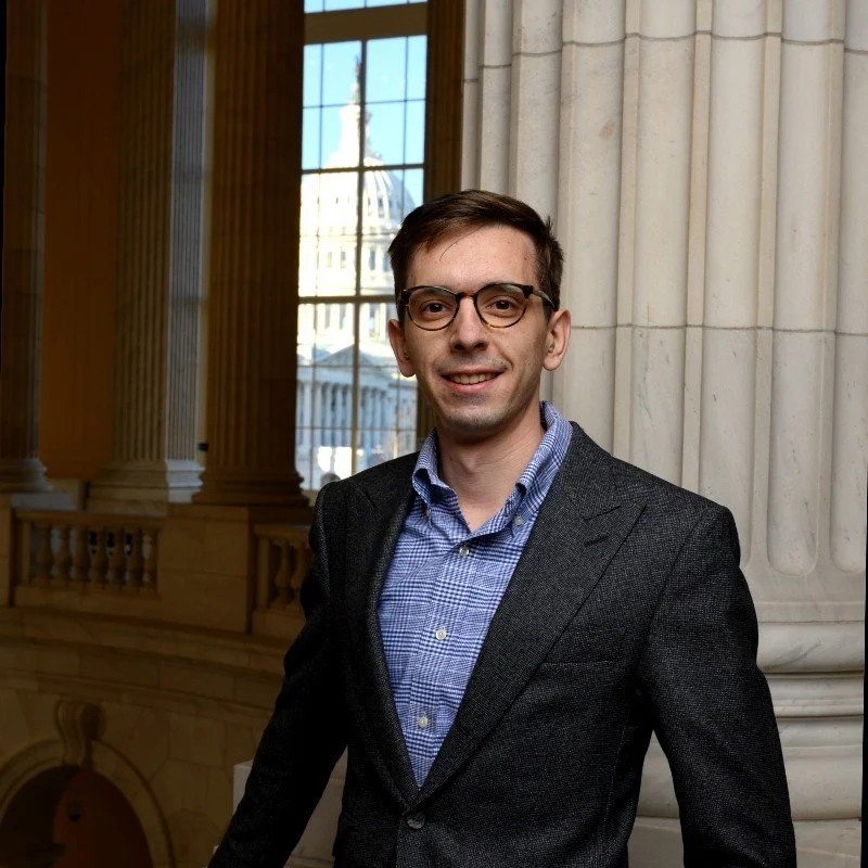A young man wearing glasses, a blue checkered shirt, and a dark blazer, standing inside a historic building with large windows and classical architectural details.