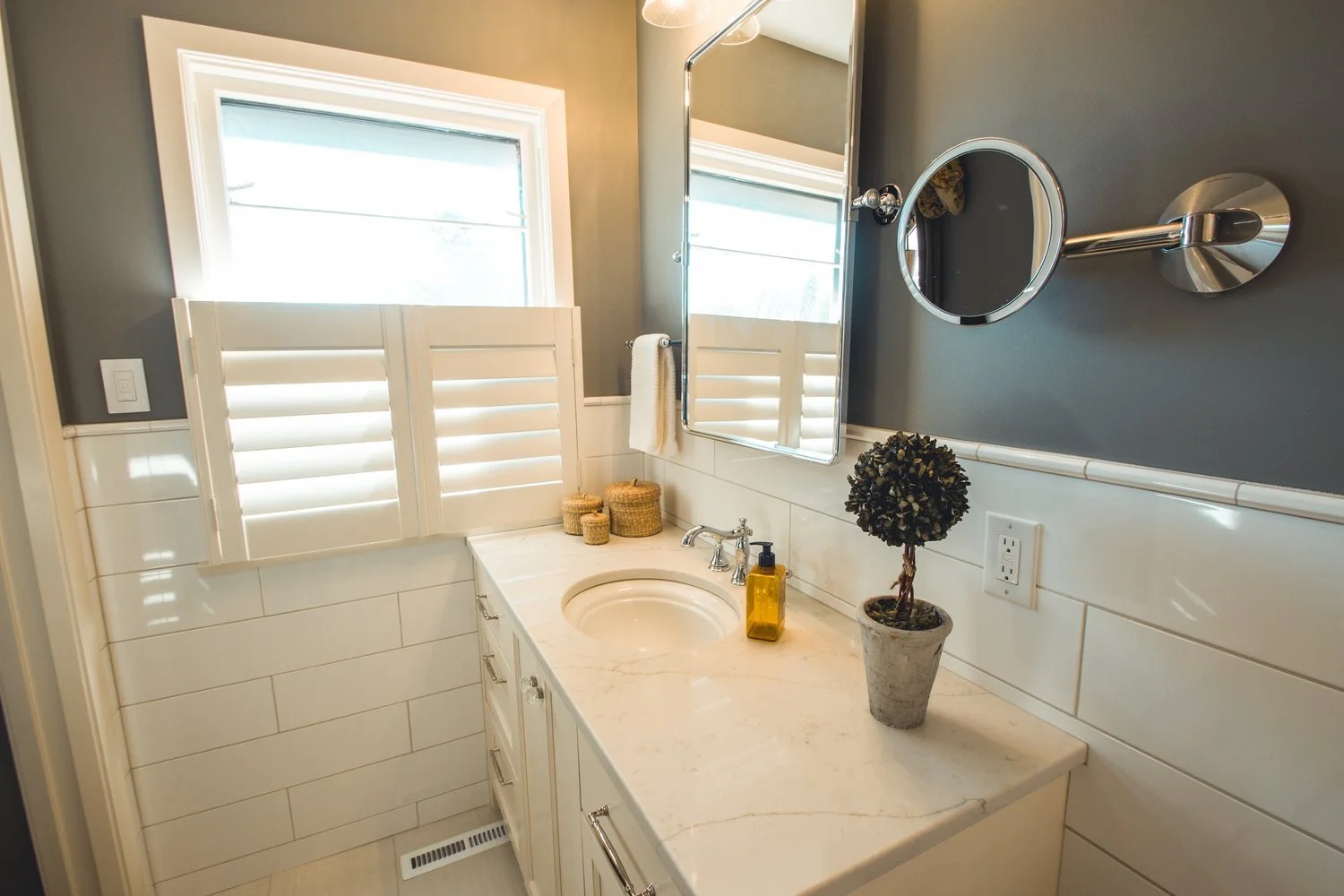 Modern bathroom in Edina with white vanity, marble countertop, round mirror, potted plant, wicker baskets, soap dispenser, and window shutters.