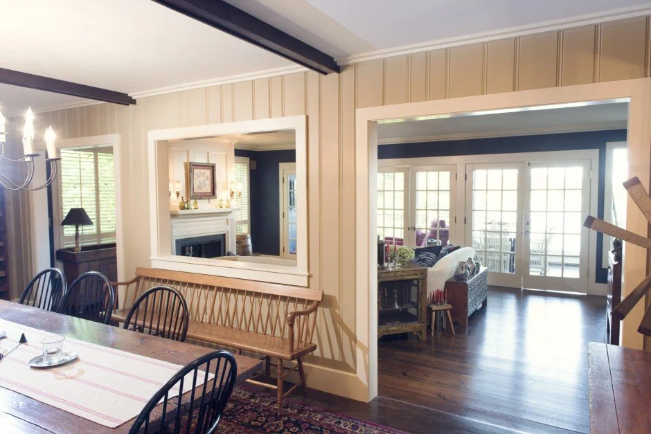 Interior of a cozy living and dining area in Savage with wooden furniture, large windows, and light-colored walls. The room features a dining table with chairs, a bench, a chandelier, and a fireplace in the adjoining room.