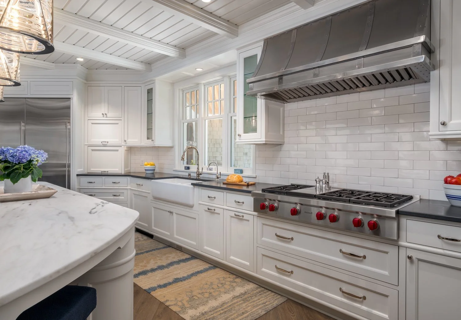 Modern kitchen with white cabinetry, stainless steel appliances, marble countertops, and a large range hood. A farmhouse sink is set beneath a window. A vase with flowers and a bowl of fruit add decorative touches.