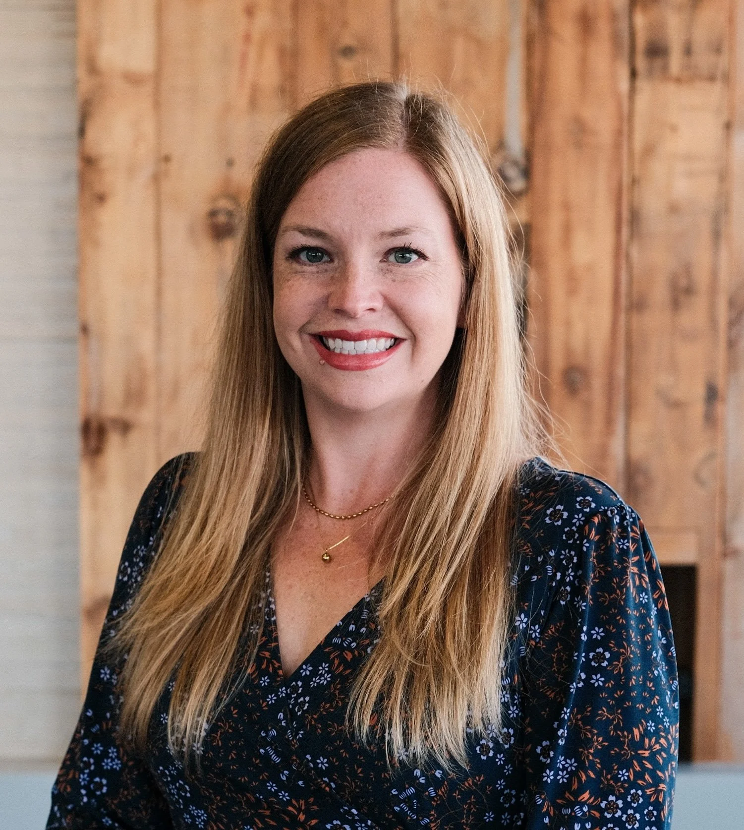 Jess with straight, long blonde hair wearing a dark floral dress, standing in front of a wooden background.