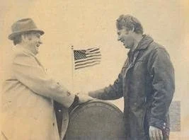 Two men shaking hands next to a barrel with a small American flag in the background.