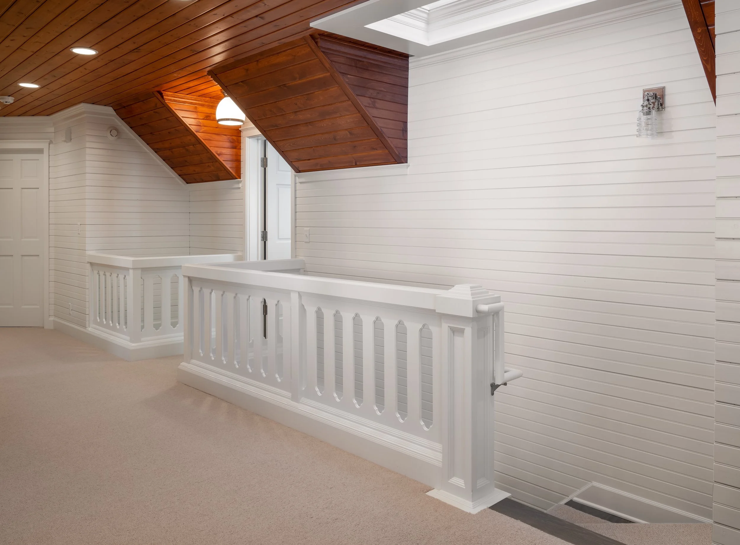 Interior hallway remodel in Wayzata with white walls and wooden ceiling, featuring a railing with decorative spindles and a carpeted floor, leading to a staircase.