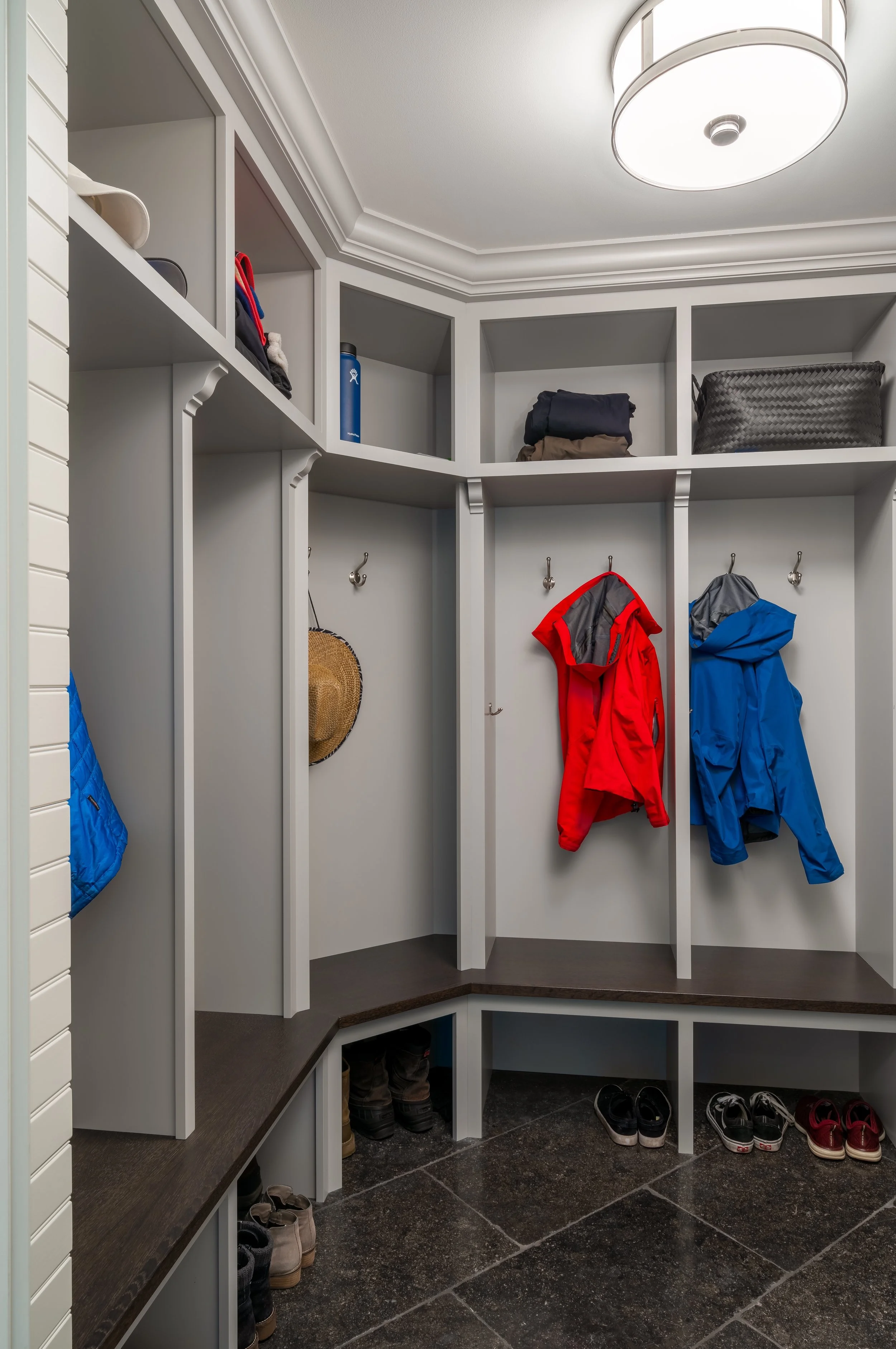 A modern mudroom in Hopkins with built-in cubbies featuring jackets, shoes, and a straw hat. Shelving above holds folded clothes and a blue water bottle. The floor is tiled, and a ceiling light fixture is visible.