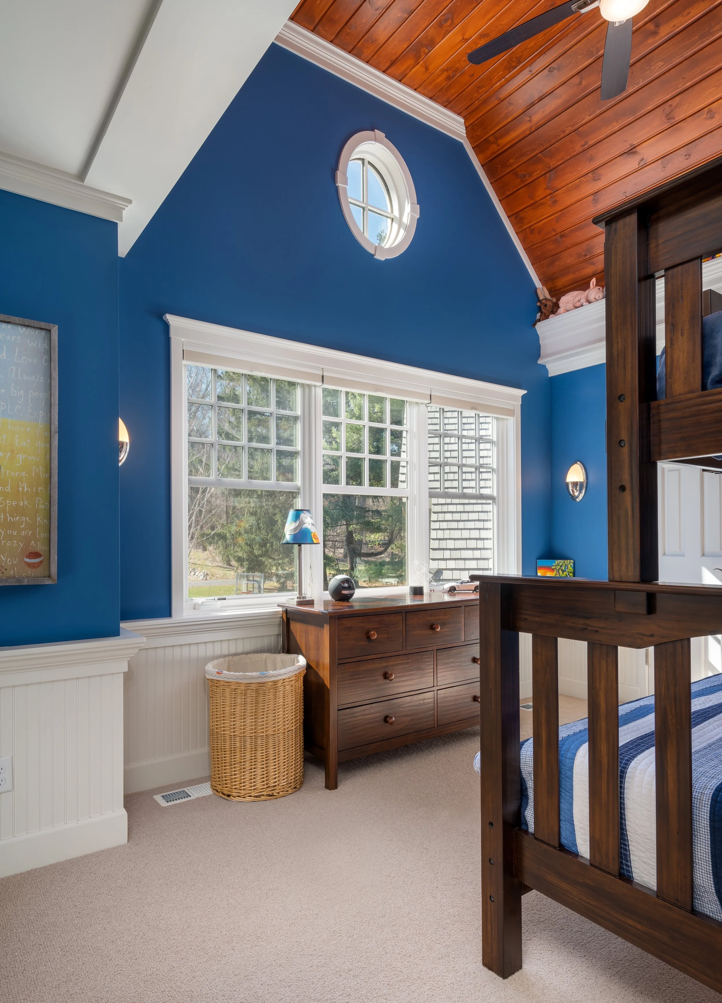 A cozy bedroom remodel in Wayzata with blue walls and a wooden bunk bed. The room features a vaulted ceiling with wood paneling, a large window with a view outside, a wooden dresser, and a wicker laundry basket. 