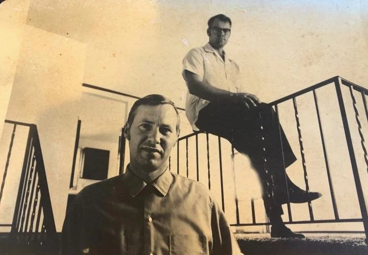 Doug Hansen in a vintage black and white photograph, one standing in the foreground and the other sitting on a railing in the background. The setting appears to be indoors with decorative railings.