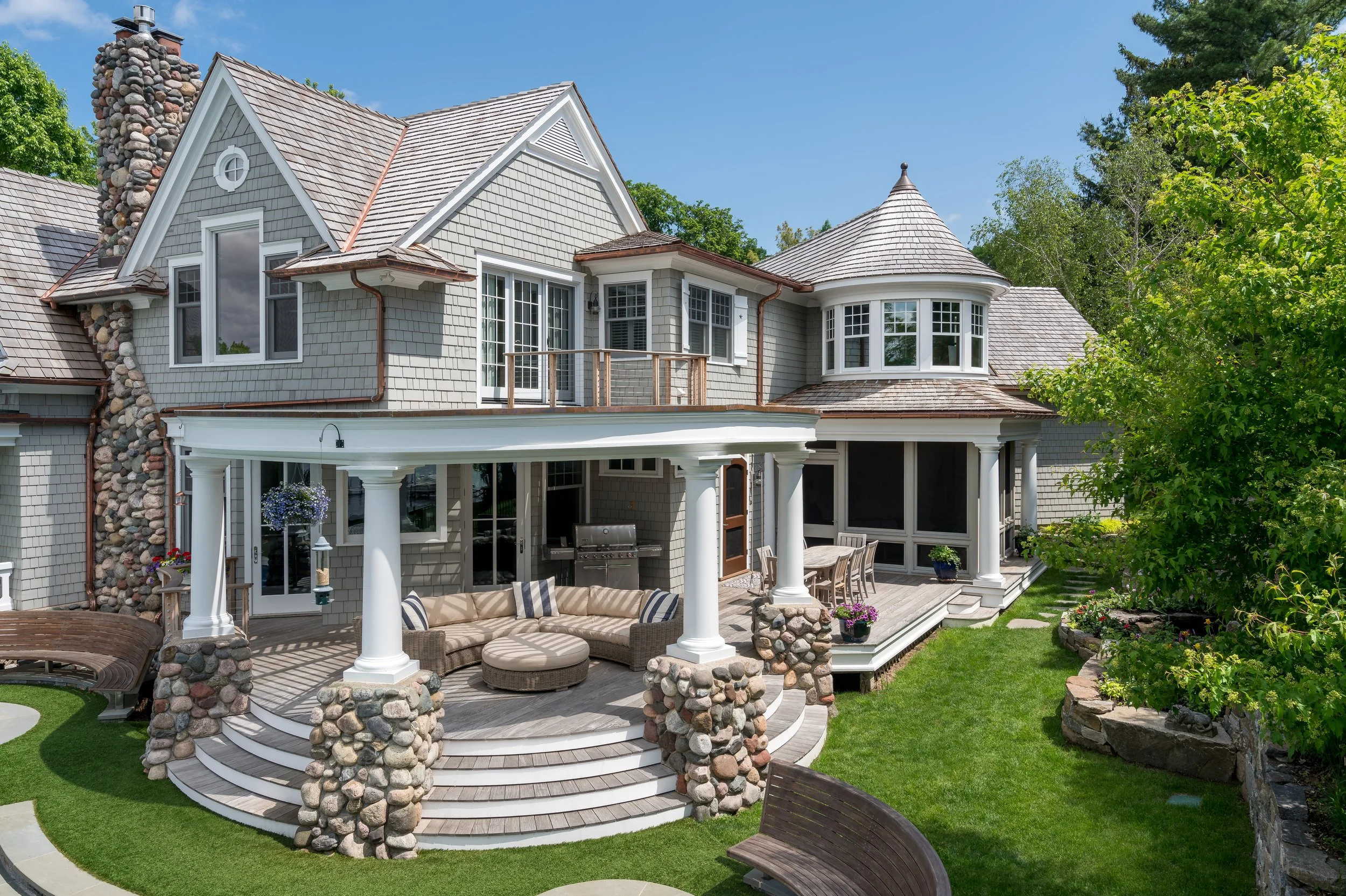 Luxury gray shingle-style house on Lake Minnetonka with circular porch and stone columns, featuring a curved sofa, outdoor dining area, and landscaped garden.