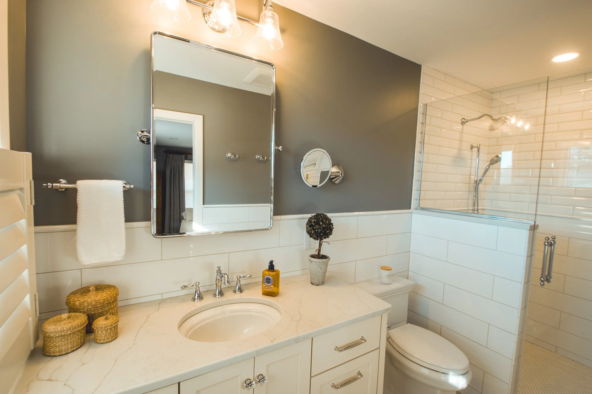 Modern bathroom in Edina with a gray and white color scheme, featuring a vanity with marble countertop, round mirror, white towels, potted plant, wicker storage baskets, soap dispenser, toilet, and glass-enclosed shower with white subway tiles.