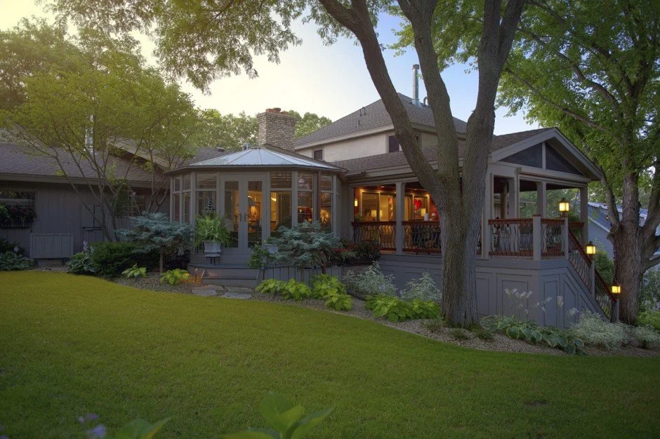Suburban house remodel in Wayzata with a glazed sunroom, porch, and well-maintained garden with trees and plants.