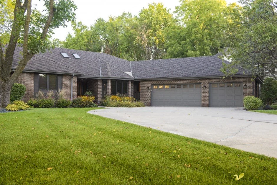 Single-story brick house  remodel in Excelsior with a large driveway and well-maintained lawn surrounded by trees.