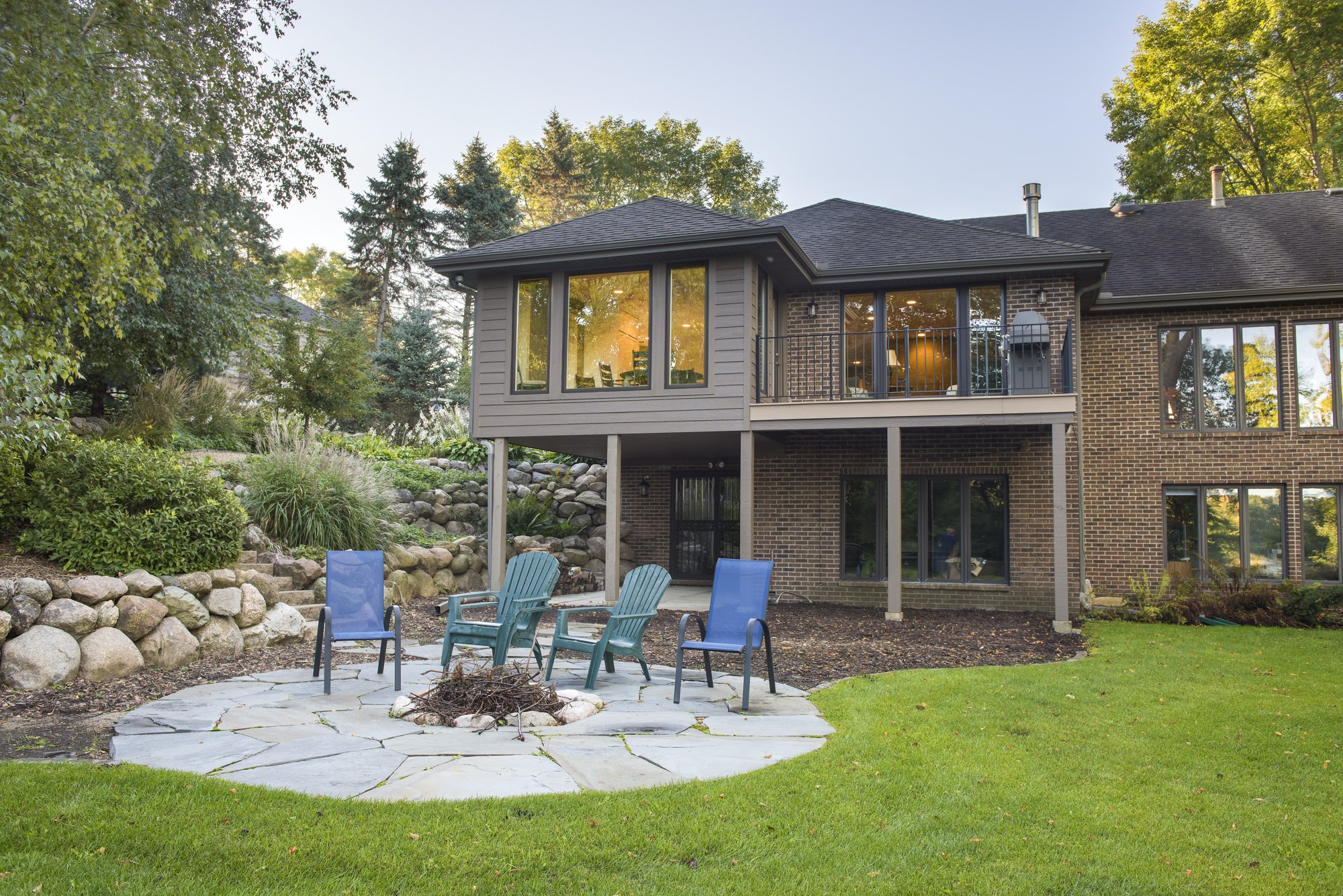 Backyard of a house addition in Excelsior with a patio area featuring Adirondack and lounge chairs, surrounded by greenery and trees.