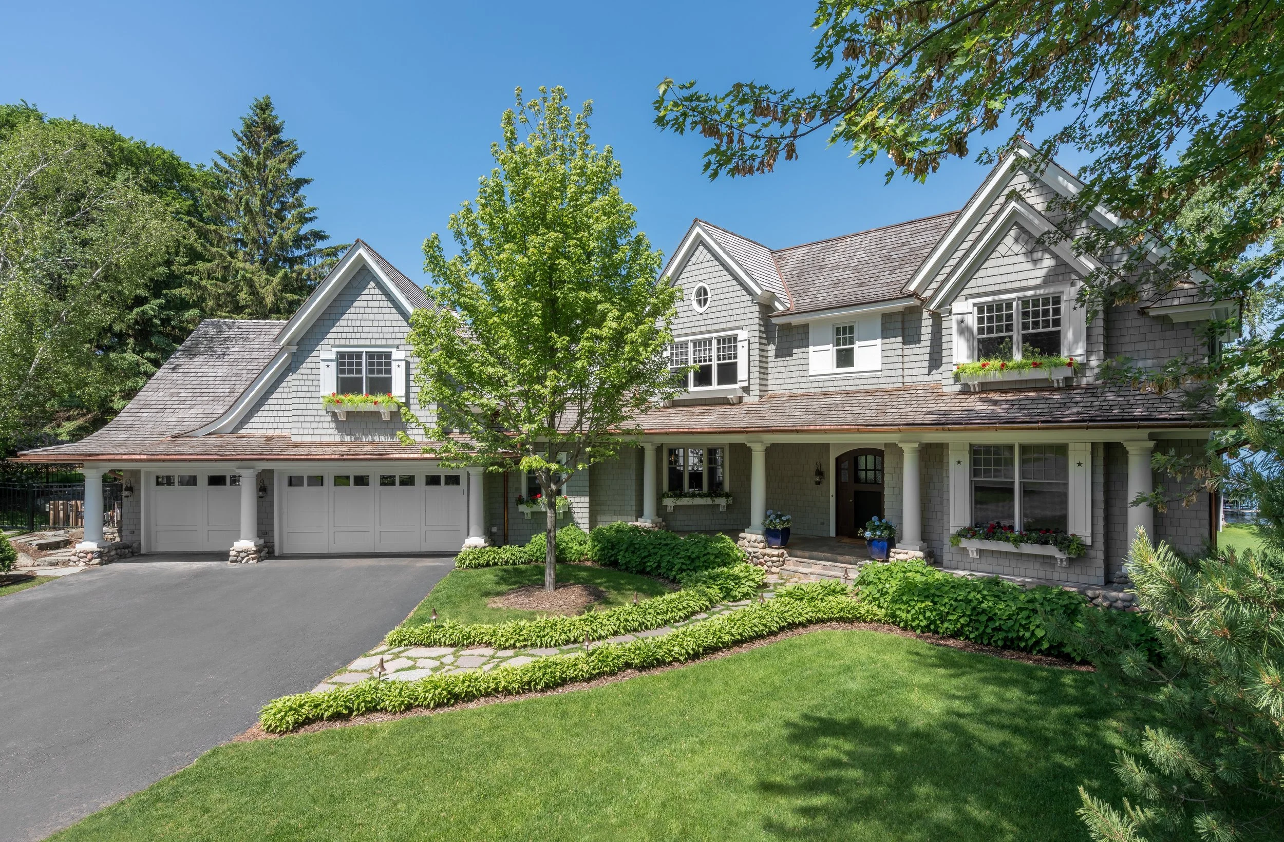 Large two-story house  remodel in Excelsior with a three-car garage, surrounded by greenery and a well-manicured lawn, featuring a front porch and windows with flower boxes.