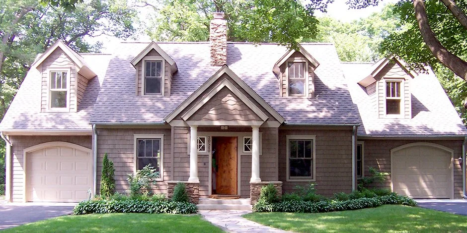A two-story house remodel in Wayzata with a gabled roof and brown siding, featuring three dormer windows and two garage doors, surrounded by trees and a manicured lawn.