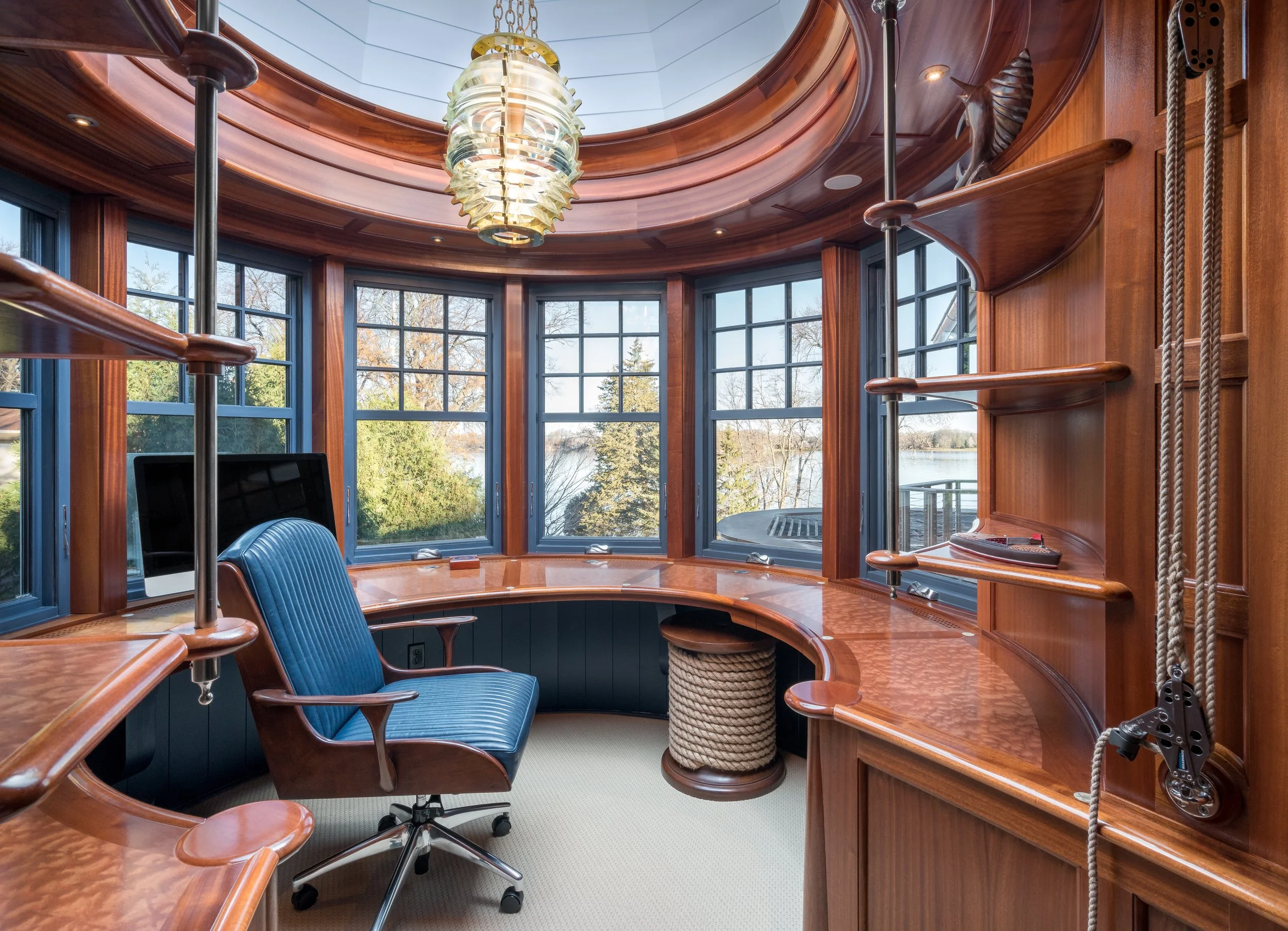 Nautical-themed home office remodel in Excelsior with wood paneling, circular desk, blue office chair, large windows, and decorative overhead light.