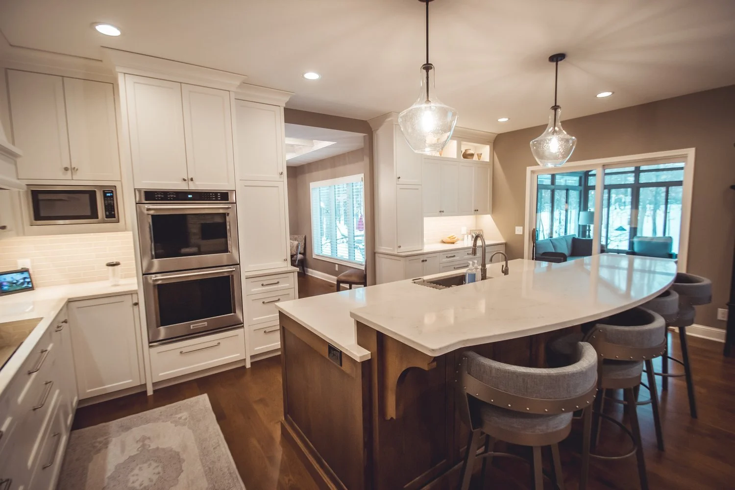 Modern kitchen remodel in Chanhassen with white cabinets, stainless steel appliances, curved island with gray stools, pendant lights, hardwood floor, and view of dining area and windows.