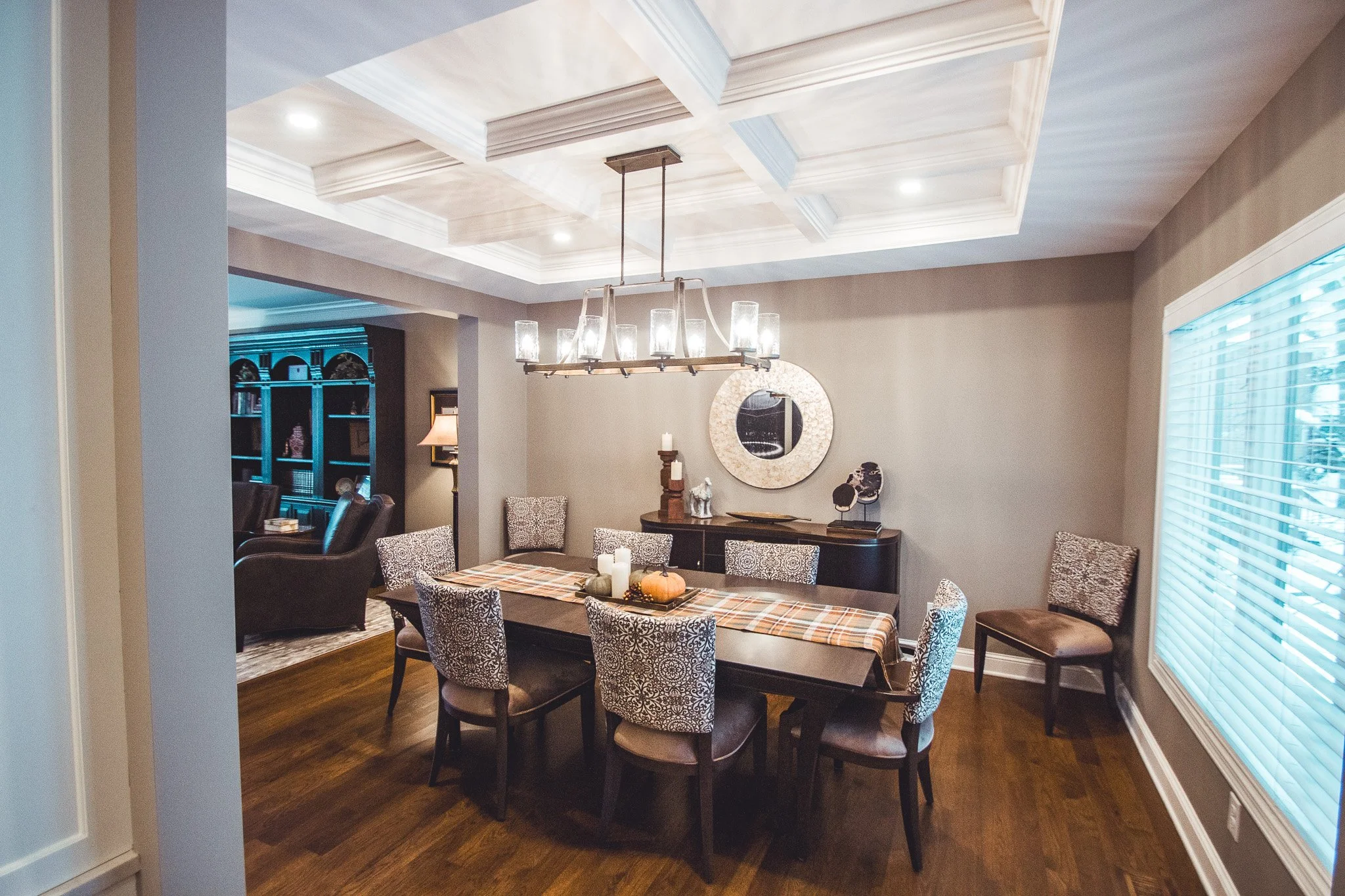 Elegant dining room remodel in Chanhassen with a large wooden table, patterned chairs, a plaid runner, and a central pumpkin decoration. Modern chandelier hanging above. Buffet with candles and a round mirror on the wall. 