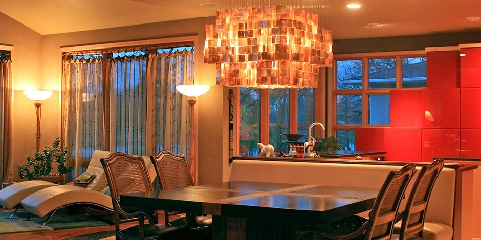 Modern living and dining area remodel in Excelsior with warm lighting, featuring a wooden dining table with chairs, a large decorative chandelier above, contemporary lounge chairs, patterned curtains, and a kitchen.