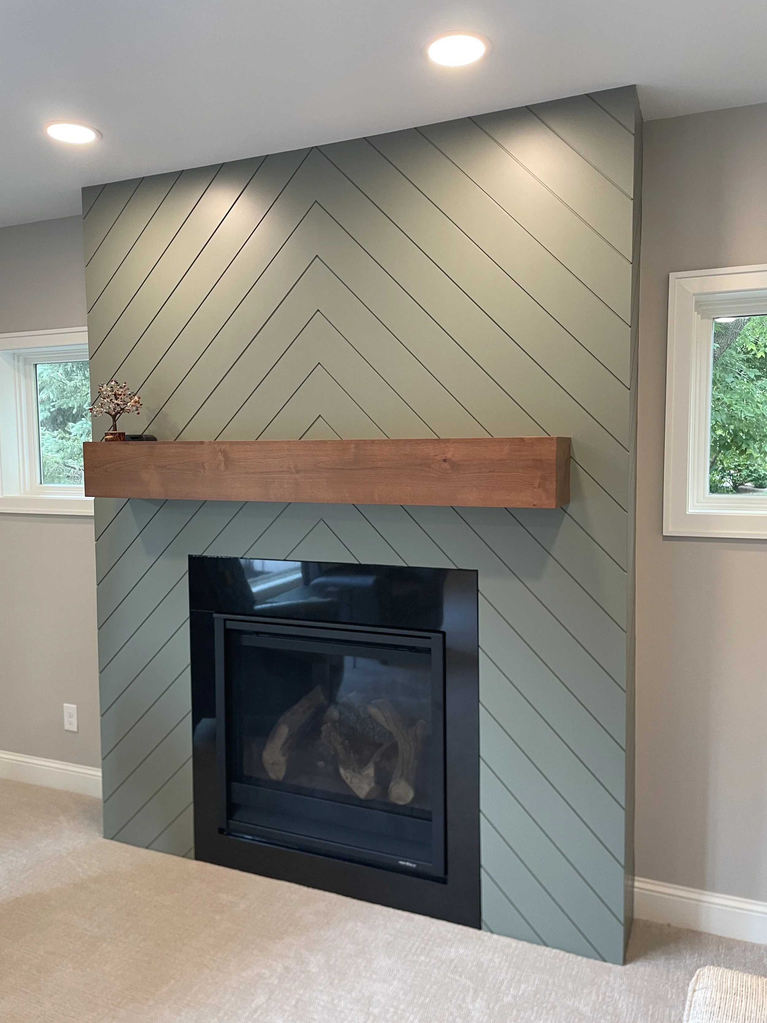 Modern fireplace remodel in Minnetonka with green herringbone patterned wall, wooden mantel, and recessed lighting in a living room.