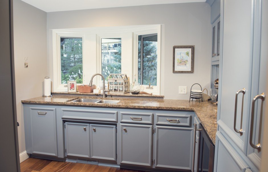 Kitchen remodel in Minnetonka with gray cabinets, brown granite countertop, double sink, paper towel holder, plant, decorative glass container, and framed picture on the wall near a window with a view of trees.