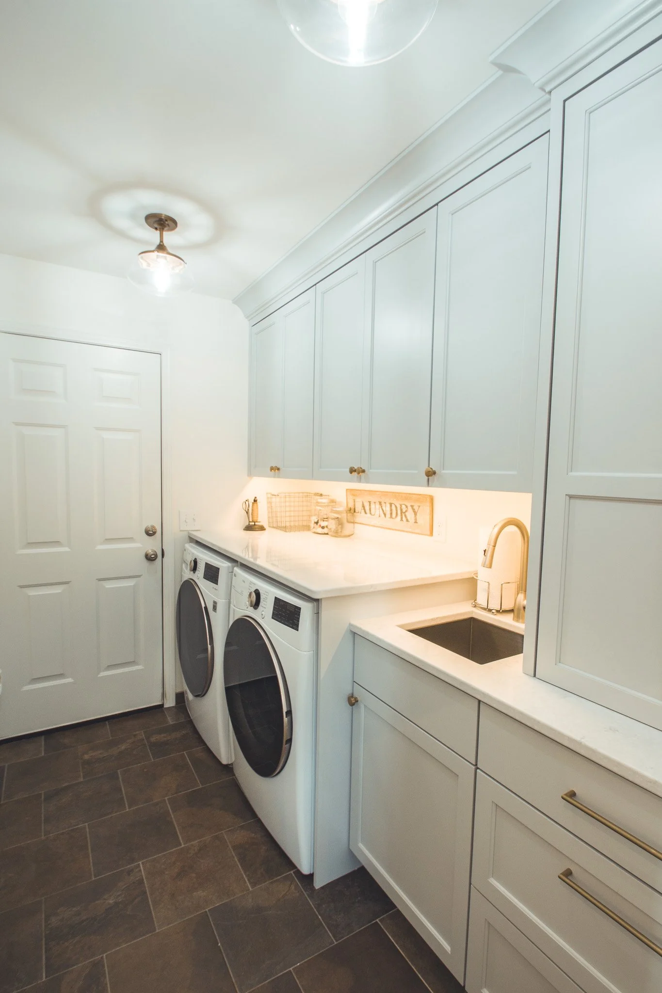 Modern laundry room remodel in Minnetonka with white cabinets, washer and dryer, and a sink, featuring dark tile flooring and overhead lighting.
