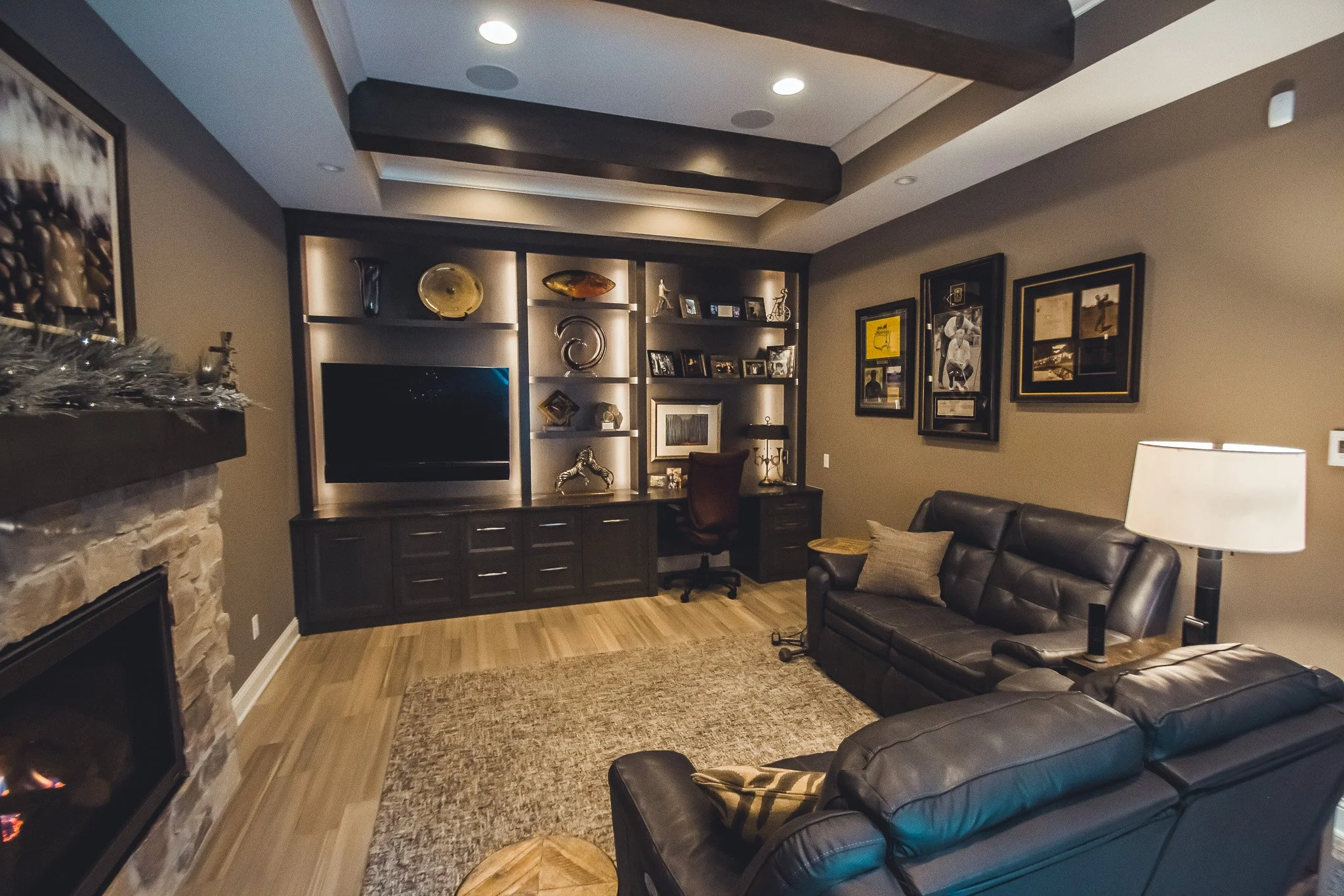 Luxurious home office remodel in Minnetonka with dark leather couch, large TV, fireplace, wooden shelves with decor, framed pictures, and a desk with an office chair.